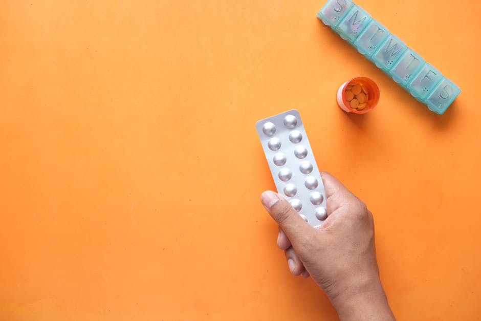 A hand holding a medicine blister near a pill organizer and orange pill bottle.