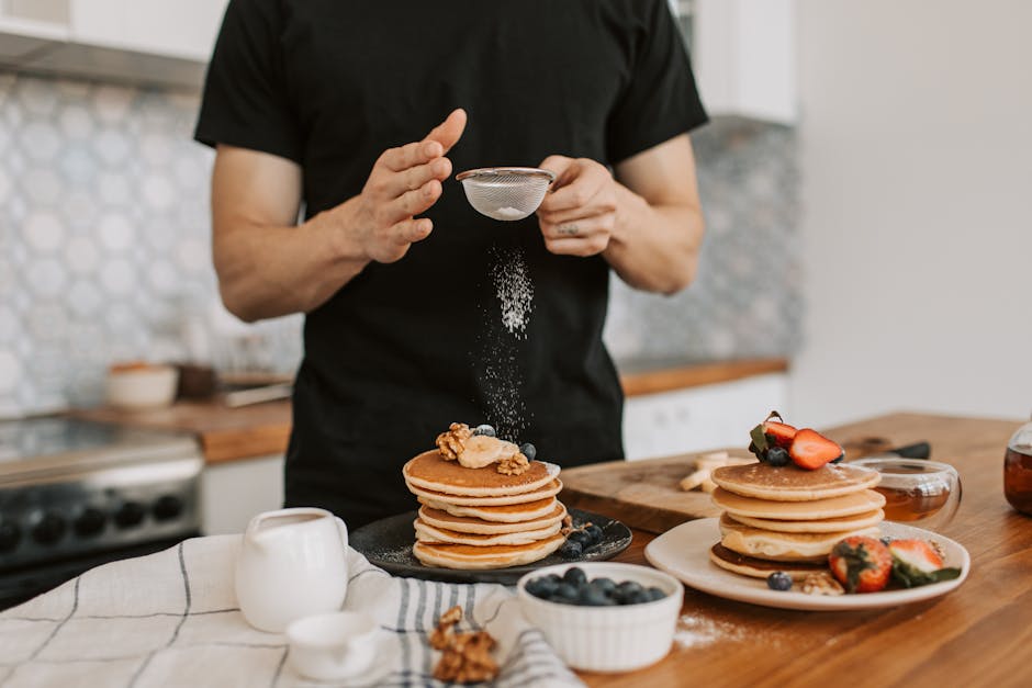 A man dusting sugar on a stack of pancakes topped with berries and nuts in a kitchen.