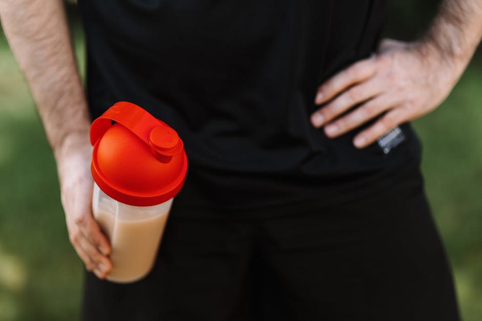 Close-up of a man holding a protein shake tumbler with a red lid outdoors, emphasizing fitness.