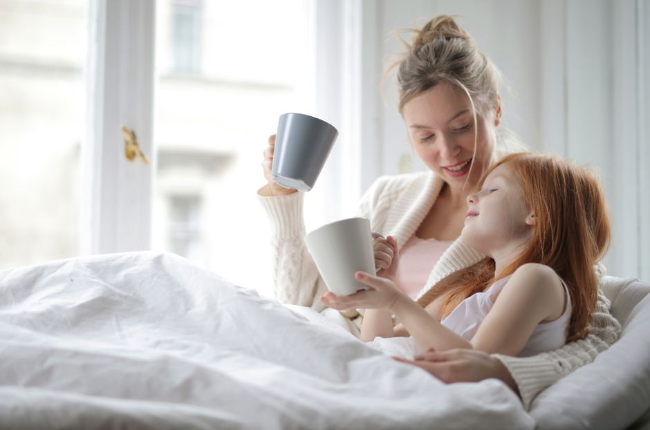 A mother and daughter sharing a warm moment with cups in bed, enjoying a cozy morning together.