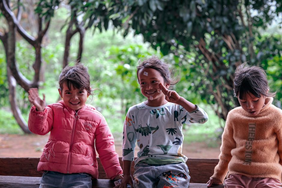 Three cheerful children sitting on a bench in a lush outdoor setting. They are smiling and interacting joyfully.