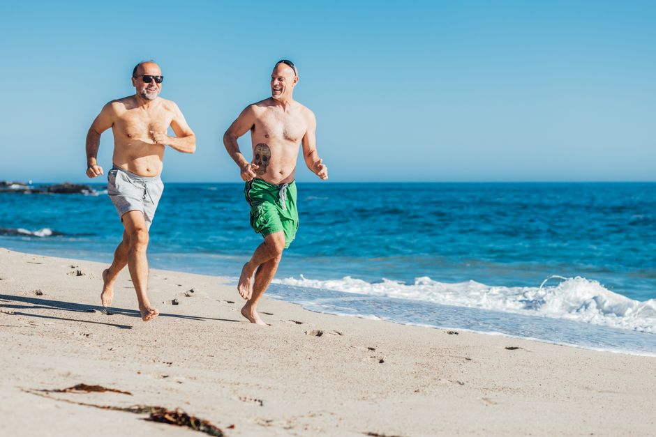 Two senior men run joyfully on a beach in summer, enjoying the ocean view under a bright blue sky.