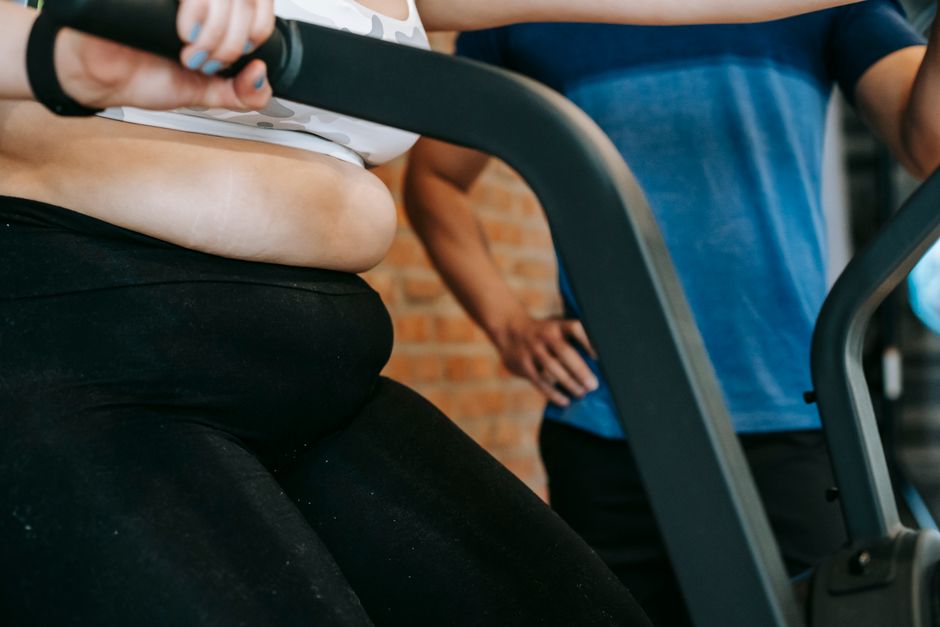 Crop faceless overweight female in sportswear doing cardio exercises on cycling equipment in gym during workout with anonymous male personal trainer