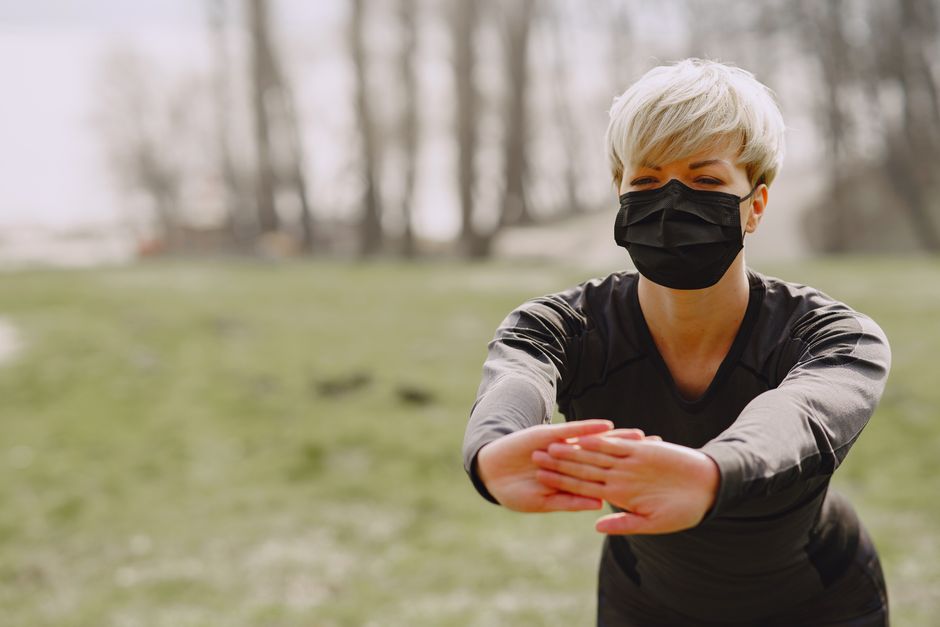 Concentrated woman in activewear and protective mask stretching arms forward and showing palms to camera while making incline in autumn park