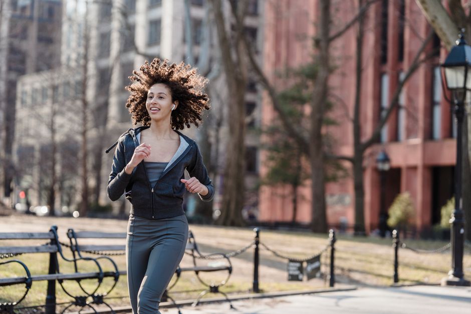 Cheerful ethnic female runner jogging on street and listening to music in wireless earphones during cardio training